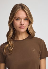 Camiseta corta de manga corta color marrón con cuello redondo. Presenta un pequeño logo en relieve en el pecho de un color contrastante. Textura suave.