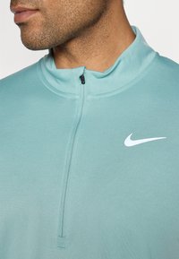 Jasnoniebieska sportowa bluzka na zamek z wysokim kołnierzem, półdługości zamkiem błyskawicznym oraz białym logo Nike z przodu. Gładka, teksturowana tkanina.