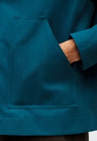 Άτομο που φοράει τιρκουάζ μπουφάν με το χέρι του μέσα στην μεγάλη μπροστινή τσέπη, δείχνοντας την υφή του υφάσματος του μπουφάν και τις λεπτομέρειες της ραφής.