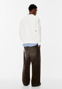 Felpa bianca con spalle scese, indossata sopra una camicia a righe blu. Pantaloni in corduroy larghi di colore marrone scuro, con tasche posteriori.