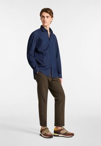 Camicia blu navy con bottoni, pantaloni marroni slim-fit e sneakers multicolori con una finitura testurizzata. La camicia presenta un colletto e dettagli sui bottoni.
