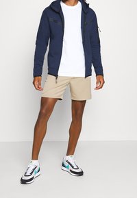 Sudadera con cremallera azul marino con detalles en negro, camiseta blanca, pantalones cortos beige, y zapatillas negro/blanco con detalles en turquesa. Tejido suave y corte atlético.