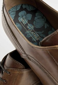 Zapato de cuero marrón con cordones negros, featuring una plantilla oscura con un patrón floral y una superficie texturizada, destacando los detalles de la artesanía.
