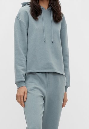Femme portant un sweat à capuche bleu clair manches longues et un pantalon de survêtement assorti, debout devant un fond uni.