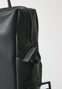 Black textured leather bag with an angular shape. Features dual black tags, a zip closure, and a prominent black clip hardware.