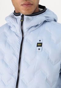 Giacca puffa azzurro chiaro con chiusura a zip, cappuccio e un motivo testurizzato. Presenta una zip nera e un'etichetta con logo vicino al petto.