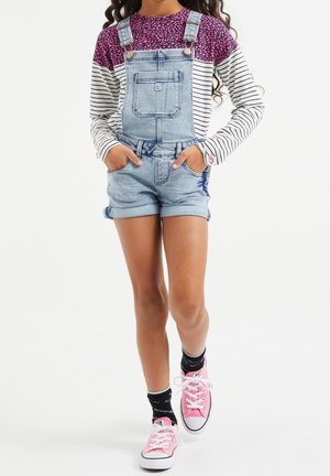 Enfant portant un t-shirt à manches longues rose avec motifs et rayures, une salopette courte en jean bleu clair, des chaussettes noires et des baskets roses, debout les mains dans les poches.