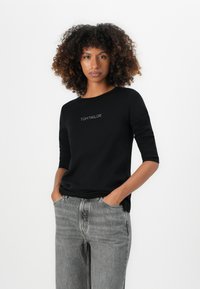 Black shirt with three-quarter sleeves, featuring the text "TOM TAILOR" in silver embroidery at the chest. Paired with gray jeans.