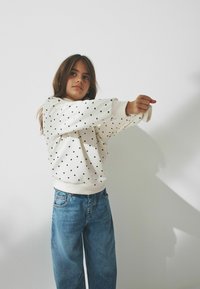 Bílá mikina s černými puntíky, žebrované manžety, kombinovaná se světle modrými džíny. Model pózuje s rozpaženýma rukama na jednobarevném pozadí.