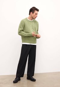 Roheline kabelikootud sviiter ribaga äärega, kantud valge särgi kohal, koos mustade laiemate pükste ja mustade kingadega, neutraalse tausta ees.