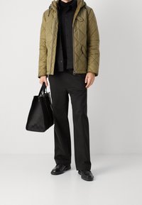 Olivgrüne gesteppte Steppjacke mit Kapuze, kombiniert mit einem schwarzen Hemd mit Knöpfen und schwarzen Marlenehosen. Hält eine schwarze strukturierte Tote Bag in der Hand.