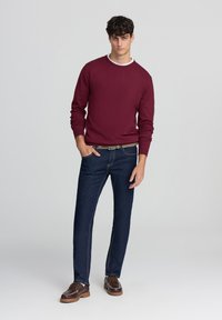 Maglione bordeaux con collo a girocollo, abbinato a jeans dritti blu scuro e mocassini marroni; texture morbida, design minimale, cuciture visibili.