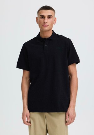 Jonge man met kort haar, gekleed in een zwart poloshirt met korte mouwen en beige broek, staande tegen een effen lichte achtergrond.