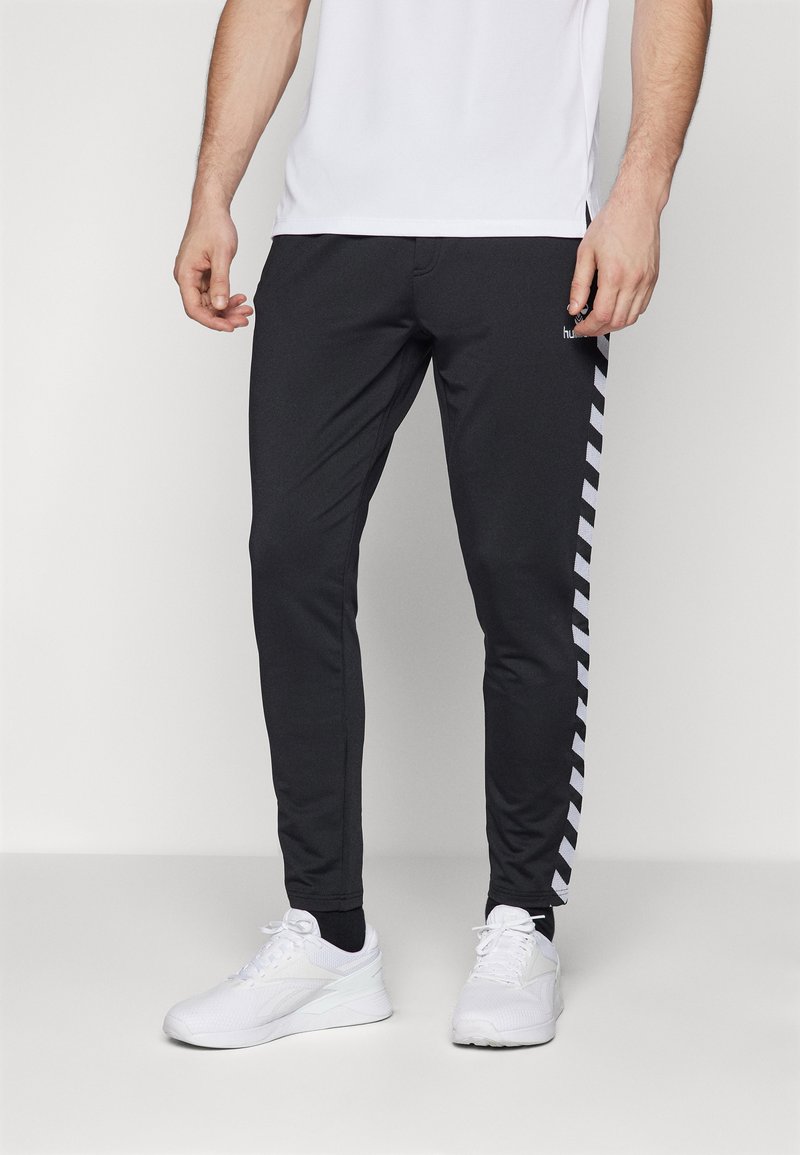 Zwarte sportbroek met een slim fit, voorzien van witte chevronstrepen langs de zijkanten. Gemaakt van rekbaar materiaal voor comfort en bewegingsvrijheid.