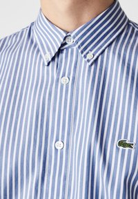 Camisa de botones a rayas azul y blanca con cuello ajustado, con un pequeño logo de cocodrilo verde y botones de plástico blancos.