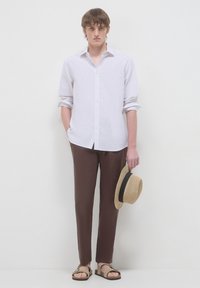 Giovane uomo con camicia bianca abbottonata, pantaloni marroni, sandali beige, che tiene un cappello di paglia con una bandiera nera, in piedi su uno sfondo bianco semplice.