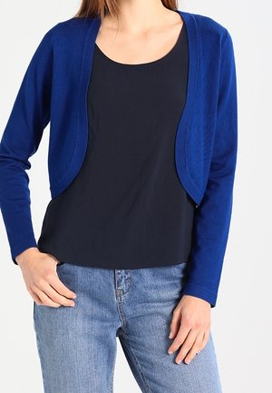Femme portant un cardigan court bleu sur un top noir, associé à un jean bleu clair, debout avec une main dans la poche et l’autre reposant le long du corps.