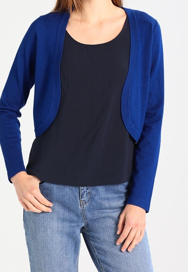 Femme portant un cardigan court bleu sur un top noir, associé à un jean bleu clair, debout avec une main dans la poche et l’autre reposant le long du corps.