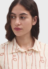 Camisa de botones color crema con un patrón de cuerda repetido en color óxido, con un collar clásico y una tela suave y texturizada.