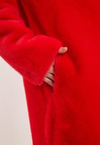 Abrigo de pelaje sintético rojo con una textura suave, mangas anchas y un bolsillo lateral visible. La tela tiene un aspecto liso y esponjoso.