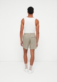 Uomo in piedi di spalle, che indossa una maglietta bianca senza maniche, pantaloni corti beige a fantasia, calzini chiari e sneakers bianche su uno sfondo neutro.