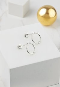 Silver hoop earrings with a sleek oval design, set on a post backing, displayed on a white surface next to a gold sphere.
