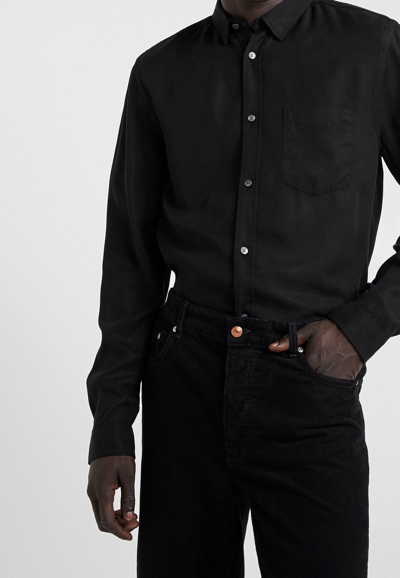 Homme portant une chemise noire boutonnée avec une poche poitrine et un pantalon noir, une main dans la poche, sur un fond uni.