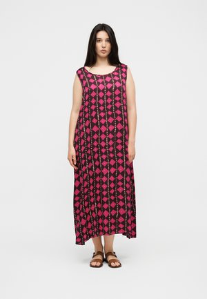 Kvinde stående iført en sort ærmeløs kjole med pink diamantmønstre og brune sandaler på en ensfarvet hvid baggrund.