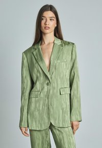 Blazer verde con texture, colletto a scollo a V, chiusura con un bottone e due tasche frontali; tessuto liscio con sottili righe.