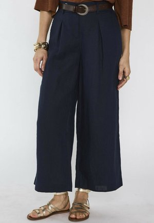 Marineblauwe wijde broek met plooien, bruine riem met decoratieve gesp, gouden sandalen met bandjes en gouden armbanden om één pols.