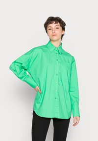Camisa verde oversized com fecho à frente, feita de tecido suave, apresentando um colarinho pontiagudo, mangas longas e um bolso no peito. Costura reforçada visível.