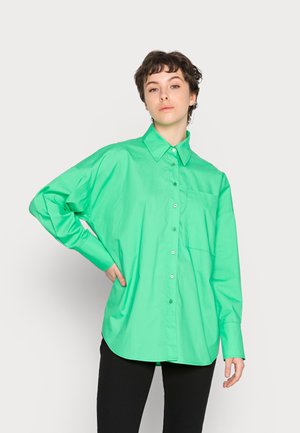 Camisa verde oversized com fecho à frente, feita de tecido suave, apresentando um colarinho pontiagudo, mangas longas e um bolso no peito. Costura reforçada visível.