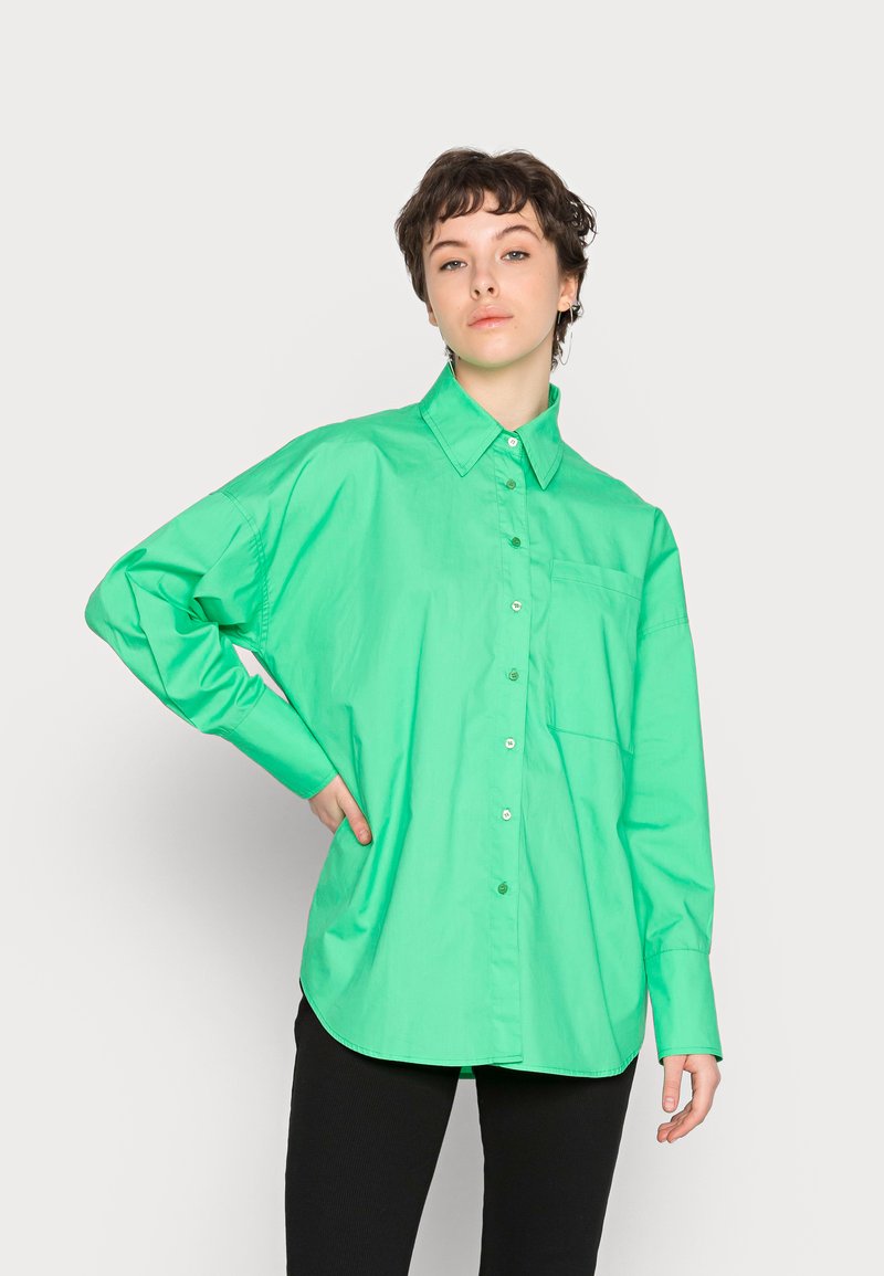Camisa verde oversized com fecho à frente, feita de tecido suave, apresentando um colarinho pontiagudo, mangas longas e um bolso no peito. Costura reforçada visível.