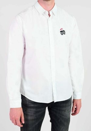 Man draagt een wit overhemd met knopen, met een klein zwart schedellogo op de linkerborst, gecombineerd met donkervervaagde jeans.