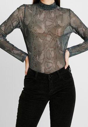 Tułów osoby ubranej w przezroczysty, długi rękaw z nadrukiem wężowym oraz czarne spodnie z wysokim stanem, ręce spoczywają na biodrach.