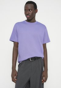 Jonge man die een lavendelkleurig T-shirt draagt, ingestopt in een hooggetailleerde grijze broek met een zwarte riem, kijkt zijwaarts tegen een witte achtergrond.