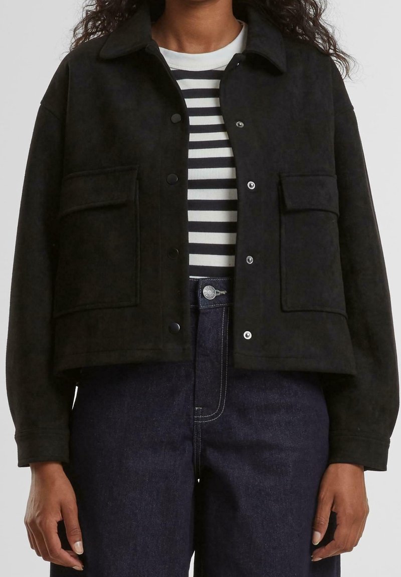 Veste courte en suédine noire avec deux poches avant, fermeture à boutons pression, et col à revers, portée sur une chemise rayée et un jean foncé.