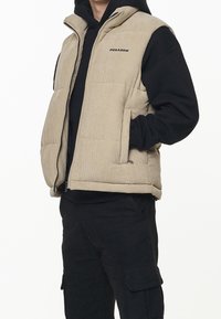 Béžová kordurová vesta s vysokým límcem, předním zipem a dvěma bočními kapsami, nošená přes černou mikinu s kapucí a černé kalhoty.