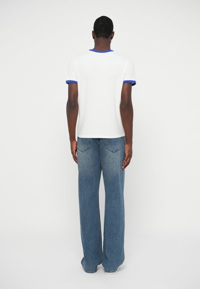 Moški stoji obrnjeno stran, oblečen v belo majico z modrim robom in ohlapne modre kavbojke, pred enobarvno ozadjem.