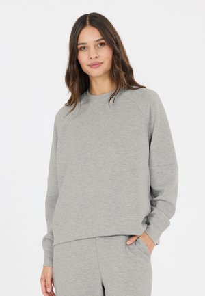 Ung kvinde med langt brunt hår iført en lysegrå sweatshirt og matchende bukser, stående med den ene hånd i lommen.
