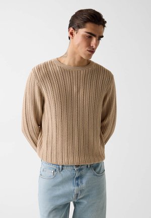 Mężczyzna w beżowym, dzianinowym swetrze o fakturze oraz jasnoniebieskich dżinsach, stojący z rękami z tyłu, patrzący w dół na bok.