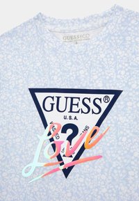 Šviesiai mėlynas organinės medvilnės marškinėliai su gėlių raštu, trikampiu mėlynai juodu logotipu su "GUESS" ir spalvingu "Meilė" dizainu viduryje.