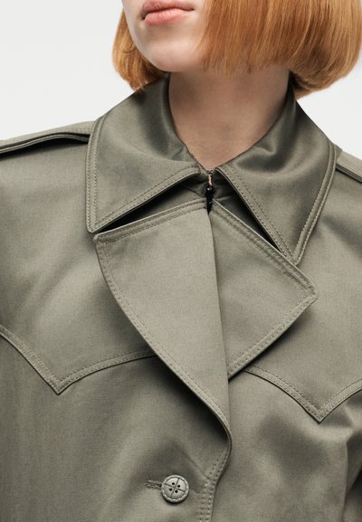 Γυναίκα με κόκκινο καρέ κούρεμα που φοράει διπλόχρωμο χακί παλτό με φαρδιές γιακάδες και λεπτομέρειες κουμπιών, εμφανίζεται από τους ώμους και πάνω.