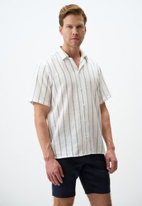 Korte mouwen linnen shirt, wit met blauwe verticale strepen, knoopsluiting aan de voorkant, puntige kraag, gecombineerd met marineblauwe shorts.