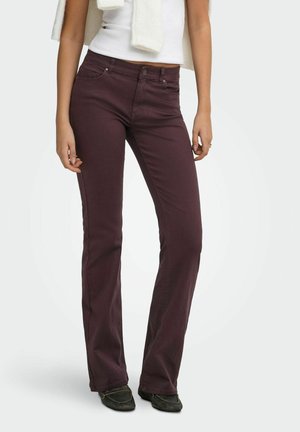 Femme portant un pantalon droit violet foncé, un top blanc sans manches, des chaussures noires et un pull clair posé sur les épaules.