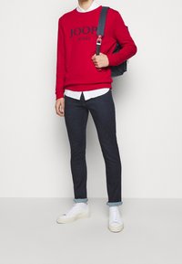 Röd sweatshirt med "JOOP JEANS" tryckt, vit skjorta med krage under, mörka jeans och vita sneakers med ljusa sulor. Svart ryggsäck.