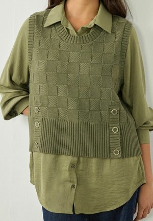 Femme portant un gilet en maille verte texturée avec des détails de boutons sur une chemise vert olive à manches longues et un jean foncé.