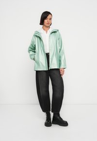 Αδιάβροχο μπουφάν σε πράσινο μέντας με κουκούλα, κλείσιμο με κουμπιά και πλαϊνές τσέπες, φορεμένο πάνω από λευκό πουκάμισο και μαύρο φαρδύ παντελόνι με μαύρες μπότες.