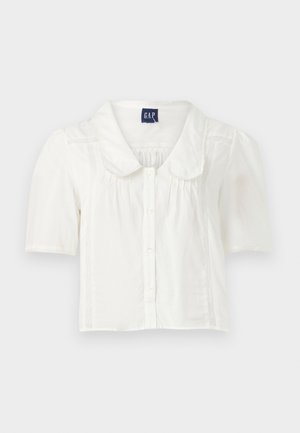 Chemisier blanc à manches courtes et col, avec fermeture à boutons, présentant de subtils plis verticaux et des accents texturés sur les côtés.