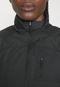 Gros plan d'une personne portant une veste noire Nike Trail avec un col haut et une poche poitrine zippée.
